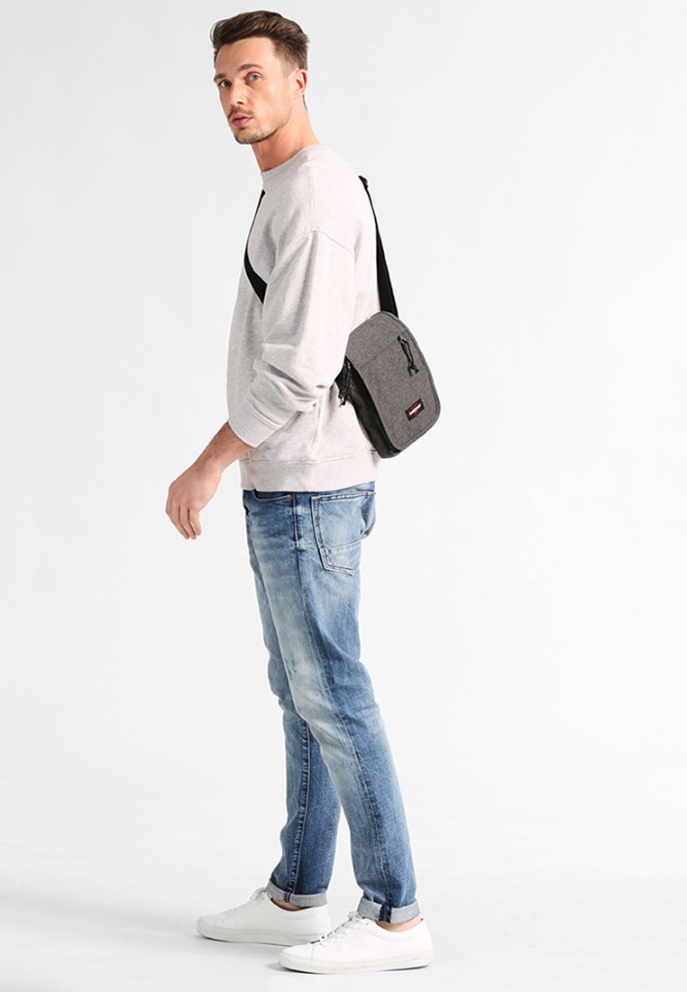 Grå budväska med dragkedjestängning, två fack och justerbar rem, bärs av modell klädd i ljusgrå sweatshirt och blekta blå jeans.