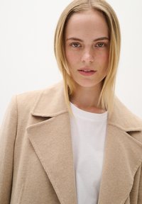 Bēšas vilnas mētelis ar iegrieztu apkakli, ar teksturētu audumu. Valkāts virs baltas kokvilnas t-krekla, atspoguļojot minimālistisku dizainu.