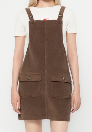 Kobieta ubrana w brązową sztruksową sukienkę na szelkach z dwoma zapinanymi na guziki przednimi kieszeniami, na białej koszuli z krótkim rękawem z małym czerwonym wzorem serca.