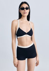 Svart ribbestrikket bikinitopp med kremfargede detaljer, med vridd front og nøkkelhullsdetalj. Matchende høytlivs ribbestrikkede svarte shorts.