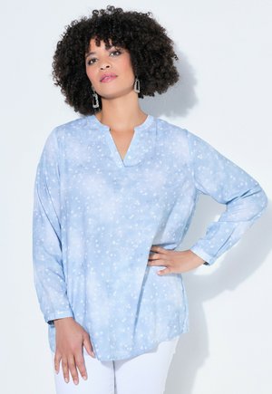 Kvinde med krøllet hår iført lyseblå bluse med lange ærmer og hvidt stjernemønster samt hvide bukser, poserer med hånden på hoften.