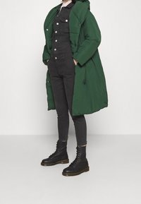 Doudoune verte avec capuche, détails de boutons et coupe oversize, associée à un jean skinny noir et des bottes noires à lacets.