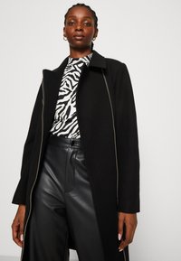 Abrigo negro con cuello de muesca, con una cremallera plateada, combinado con una blusa de estampado de cebra en blanco y negro y pantalones de piel de tiro alto.