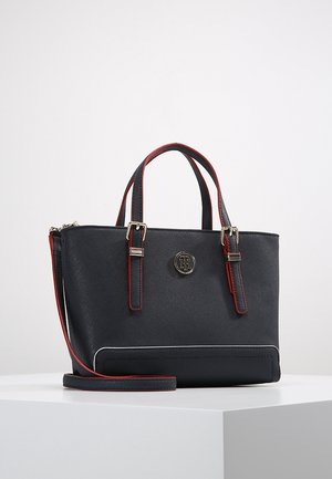 Sac à main noir texturé avec doubles poignées supérieures, bordure rouge, bandoulière amovible, boucles argentées et un emblème rond en métal avec logo sur le devant.