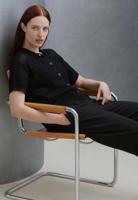 Schwarz gestricktes Polohemd mit kurzen Ärmeln und Knopfleiste, kombiniert mit schwarzen Hosen. Das Model sitzt auf einem Stuhl aus Chrom und Holz.