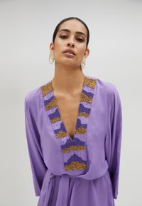 Ljubičasta haljina s dubokim V-izrezom i dekorativnim beaded ovratnikom s uzorcima u zlatnoj, plavoj i ljubičastoj boji; mekani materijal i lepršava silueta.
