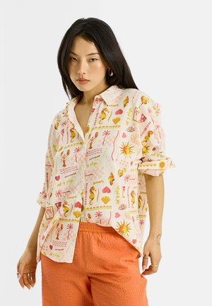 Femme aux longs cheveux foncés portant une chemise crème avec des motifs colorés de la mer et du soleil et un pantalon texturé orange, debout sur un fond blanc.
