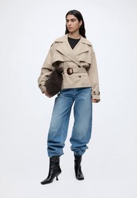 Γυναίκα με μπεζ καμπαρντίνα με ζώνη, φαρδιά ανοιχτό μπλε τζιν περασμένα μέσα σε μαύρα μποτάκια με τακούνι, κρατώντας σκοτεινή καφέ τσάντα στον βραχίωνα.