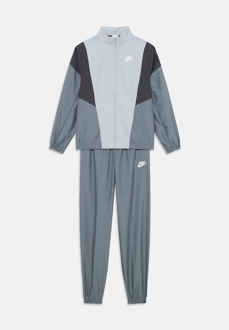 Sivi sportski trenirka koja se sastoji od jakne s patentnim zatvaračem s crnim detaljima, dugim rukavima i odgovarajućih hlača s elastičnim manžetama i Nike logom.