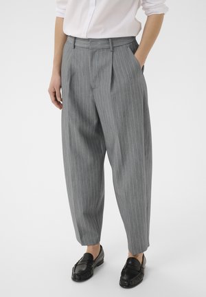 Personne portant un pantalon large à rayures grises, une chemise blanche et des mocassins noirs, debout avec une main dans la poche.