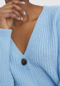 Osoba w jasnoniebieskim swetrze z dzianiny, z pojedynczym ciemnym guzikiem, z palcami blisko brody i pasującym jasnoniebieskim manicurem.