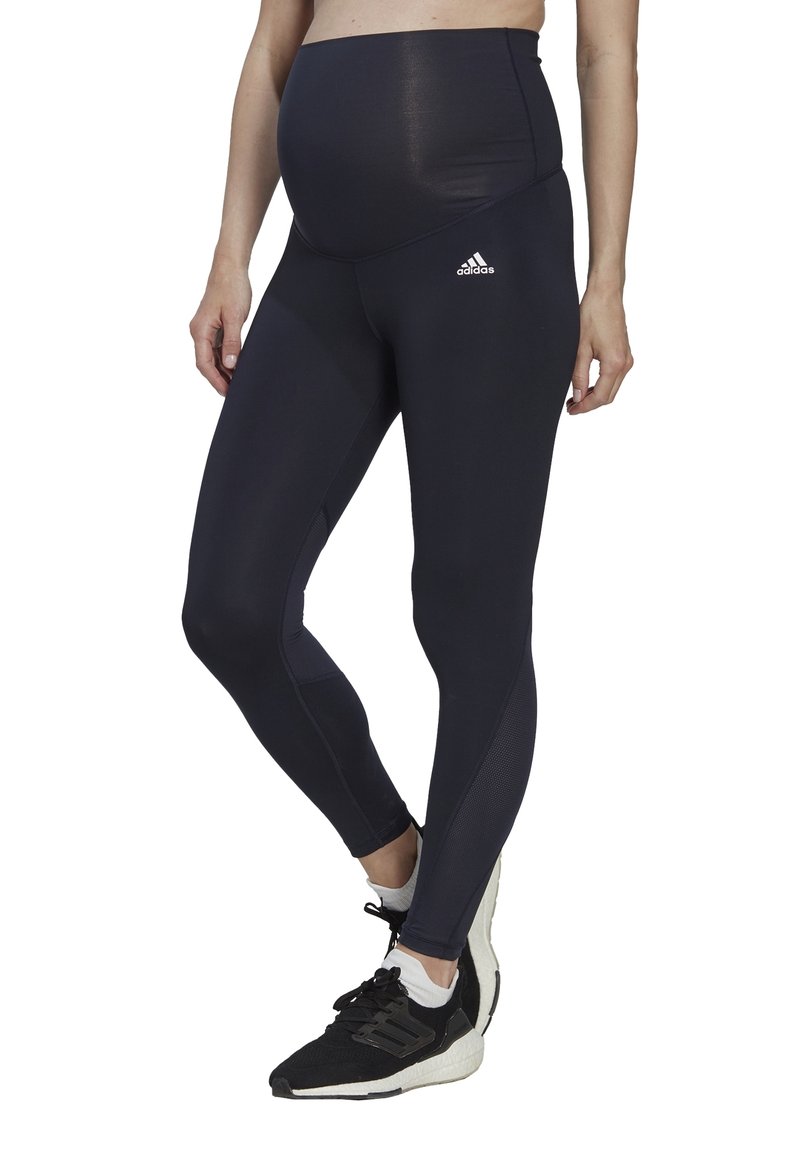 Leggings da maternità neri con vita alta, caratterizzati da una consistenza liscia e un branding discreto. Abbinati a scarpe da ginnastica nere.