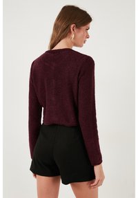 Burgundská, zkrácená, chlupatá pletená mikina s dlouhými rukávy; nošena přes černé, dokonale padnoucí šortky. Jednoduchý, zdobením neobohacený design s texturovaným povrchem.