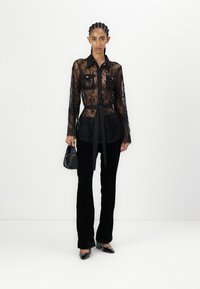 Camicia in pizzo nero con maniche lunghe, tasche frontali e una cintura in vita. Abbinata a pantaloni neri e una borsa a tracolla di piccole dimensioni. Tacchi neri lucidi completano il look.