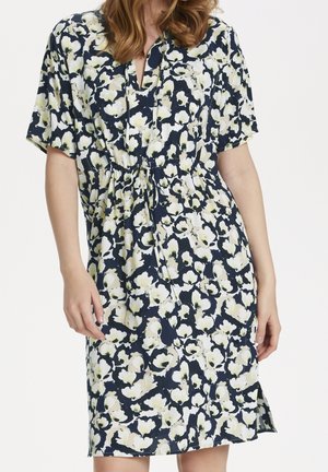 Kvinde iført en knælang kjole med korte ærmer, mørkeblå og hvidt blomsterprint og snørre talje.