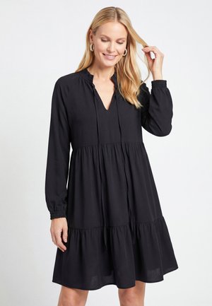 Frau mit langen blonden Haaren, die ein lockeres schwarzes langärmeliges Kleid mit gerafften Bündchen und einem Stufenrock trägt, berührt ihr Haar und lächelt.