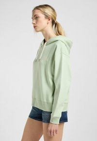 Sudadera con capucha de color verde claro y corte relajado, con un logo en la parte frontal y cordones. Combinada con pantalones cortos de mezclilla, mostrando puños acanalados.