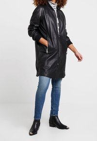 Zwarte leren lange jas met zijzakken, ritssluiting aan de voorkant, geribde manchetten, gedragen over een lichtgrijze turtleneck, gecombineerd met blauwe spijkerbroeken en enkellaarsjes.
