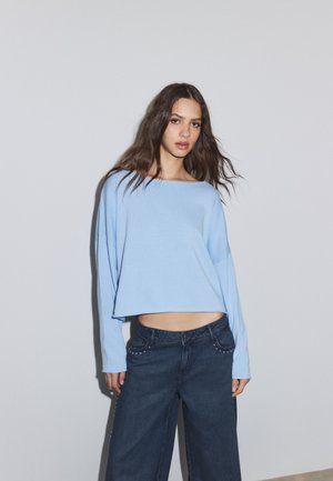 Young woman with long brown hair wearing a light blue loose long-sleeve top and dark wide-leg jeans, standing against a plain gray wall.