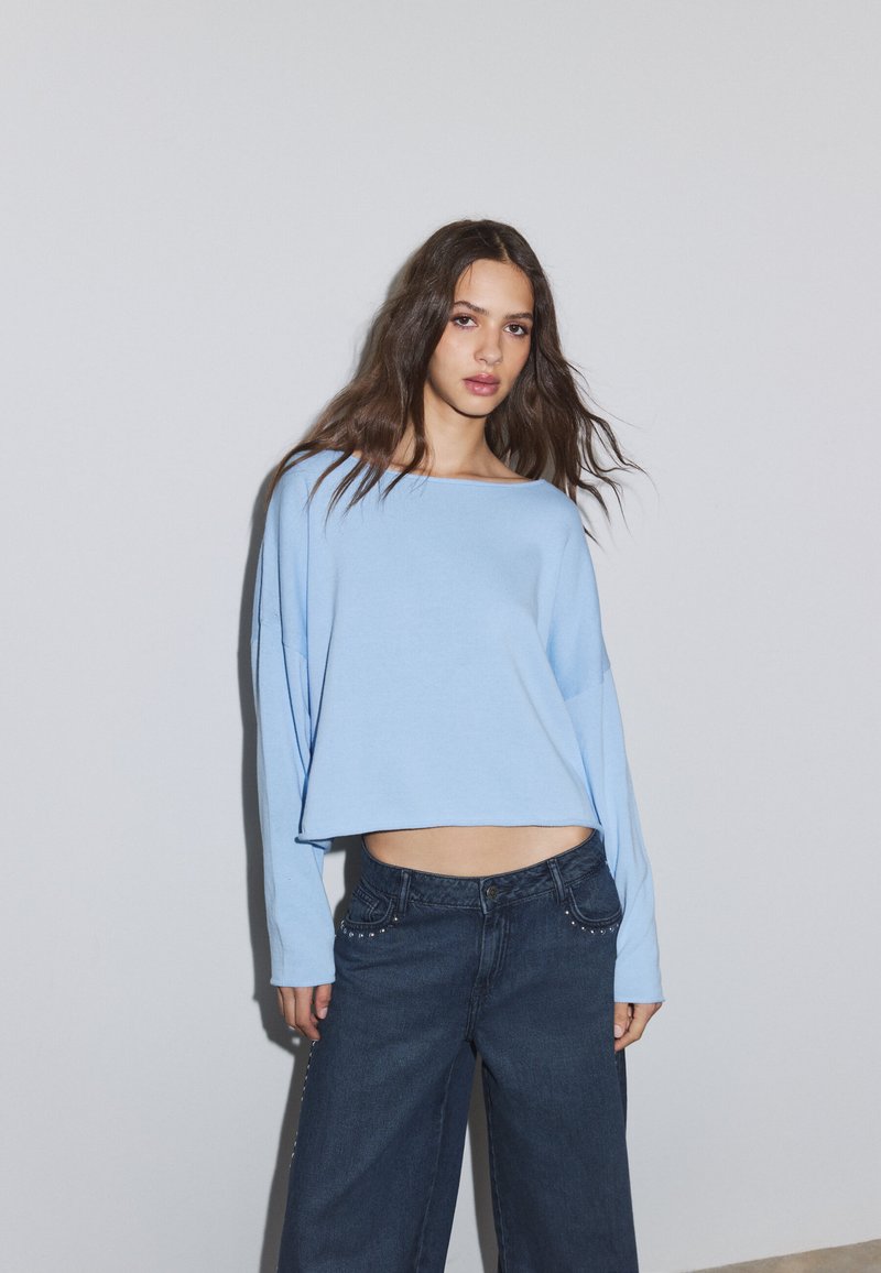 Jeune femme aux longs cheveux bruns portant un haut ample à manches longues bleu clair et un jean large et foncé, debout devant un mur gris uni.
