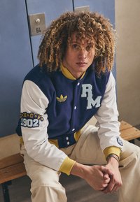 Mornarsko modra in bela varsity jakna z rumenimi detajli, z vezenim "RM" in "1902" na rokavu, oblečena preko smetanastih hlač.
