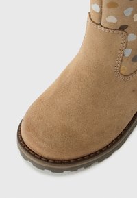 Bottine beige en suède avec un bout arrondi, des accents cousus et une semelle en caoutchouc texturé. La section en tissu à motif présente des pois discrets.