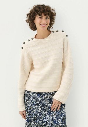 Pull à rayures crème avec un tricot texturé, un col rond et des boutons noirs décoratifs sur les épaules, associé à une jupe fleurie.