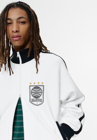 Ung person med lange dreadlocks bærer en hvid zip-up jakke med en sort krave og globe emblem med gyldne stjerner på brystet.