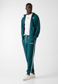 Turkooise trainingspak met witte zijstrepen, voorzien van steekzakken met rits; gecombineerd met wit T-shirt met rood "GUESS"-logo en witte sneakers.