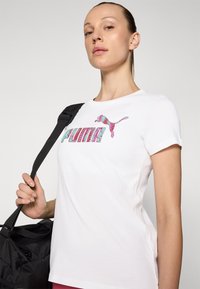 Camiseta blanca de algodón con un colorido logo de "PUMA" y un logo de gato en rosa y turquesa. El modelo sostiene una bolsa negra con una correa para el hombro.