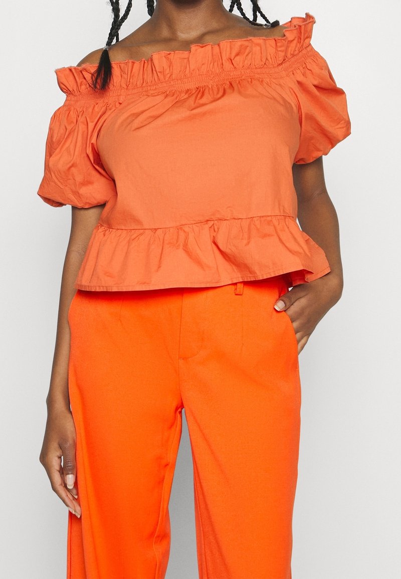 Orange off-shoulder topp med volangkrage och korta puffärmar, med en peplumslutning. Matchas med matchande orange byxor.