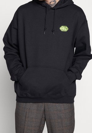 Felpa nera con cappuccio, tasca frontale e logo verde "ENNACE" sul petto, indossata con pantaloni grigi a quadri, mani infilate nelle tasche.