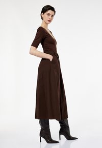 Mujer de pie con las manos en los bolsillos, vistiendo un vestido marrón con escote en V y botas de tacón negro contra un fondo blanco.