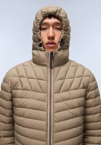 Beige puffjakke med høj hætte og snørebånd, diamantquiltet mønster og frontlynlås. Glat tekstur med en tætsiddende silhuet.