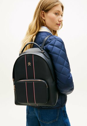 Blonde vrouw met een blauw doorgestikt jack en spijkerbroek, draagt een zwarte leren rugzak met rode en witte strepen en een gouden logo op de achterkant.