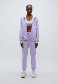 Φούτερ με φερμουάρ ανοιχτό και καπαρντίνα σε ανοιχτό μωβ χρώμα με μπροστινές τσέπες και κορδόνι, σε συνδυασμό με παντελόνι φόρμας στο ίδιο χρώμα. Το σύνολο ολοκληρώνεται με λευκά αθλητικά παπούτσια.