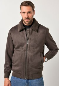 Bruine suède bomberjack met een gestructureerde bruine bontkraag, ritssluiting aan de voorkant en geribde manchetten, gestyled over een zwart shirt en blauwe jeans.