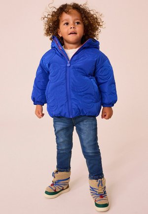 Saphirblaue gesteppte Jacke mit Kapuze, die strukturierte Smiley-Gesichtsmuster und einen Frontreißverschluss aufweist. Getragen mit Jeans und cremefarbenen Stiefeln.