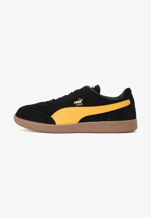 Sneakers en suède noir avec une bande en cuir jaune, semelle en caoutchouc marron et détail du logo doré sur le côté. Design bas avec des lacets.