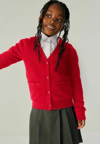 Roter gestrickter Cardigan mit langen Ärmeln, V-Ausschnitt, zwei Fronttaschen und rosa Knöpfen, getragen über einem weißen Hemd mit Kragen und einem grauen Rock.