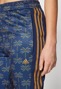 Mörkblå byxor med ett palmtoppmönster i guld och blått. Har guldstrimmor längs sidorna och en Adidas-logotyp framtill.