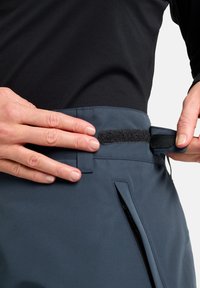 Blauer Stoffbund mit Klettverschlussakzenten und Reißverschlussdetails. Hände passen den Bund an und zeigen das strukturierte Material und das funktionale Design.