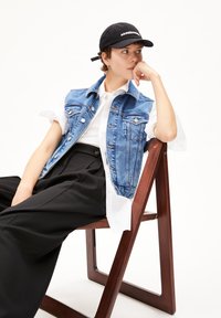 Die Jeansweste besteht aus blauem Stoff mit leichter Waschung, silberner Hardware und wird über einem weißen Knopfhemd getragen, kombiniert mit weiten schwarzen Hosen.