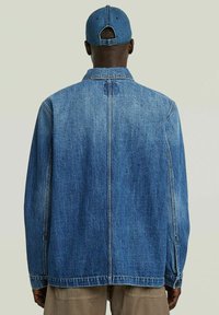 Uomo che indossa una giacca di jeans blu e un cappello di jeans blu coordinato, in piedi con le spalle alla telecamera su uno sfondo neutro.