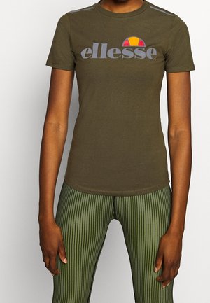 Personne portant un t-shirt vert olive avec le logo ellesse et un pantalon ajusté rayé verticalement vert et noir, debout avec les bras détendus.