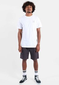 T-shirt en coton blanc avec un petit motif multicolore sur le côté gauche de la poitrine. Associé à un short cargo foncé et des baskets noires avec des accents blancs.
