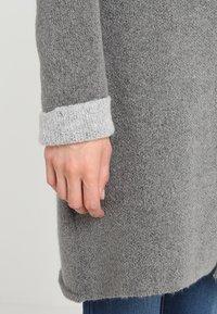 Grå ulltröja med en mjuk textur, med en ljusgrå ribbad mudd vid handleden. Handen vilar mot tr öjans sida.