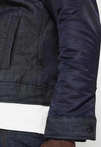 Marinblå jacka med denimkropp och en blank, slät satinärm till vänster. Inkluderar en vit ribbad fåll och en märkesetikett på fickan.