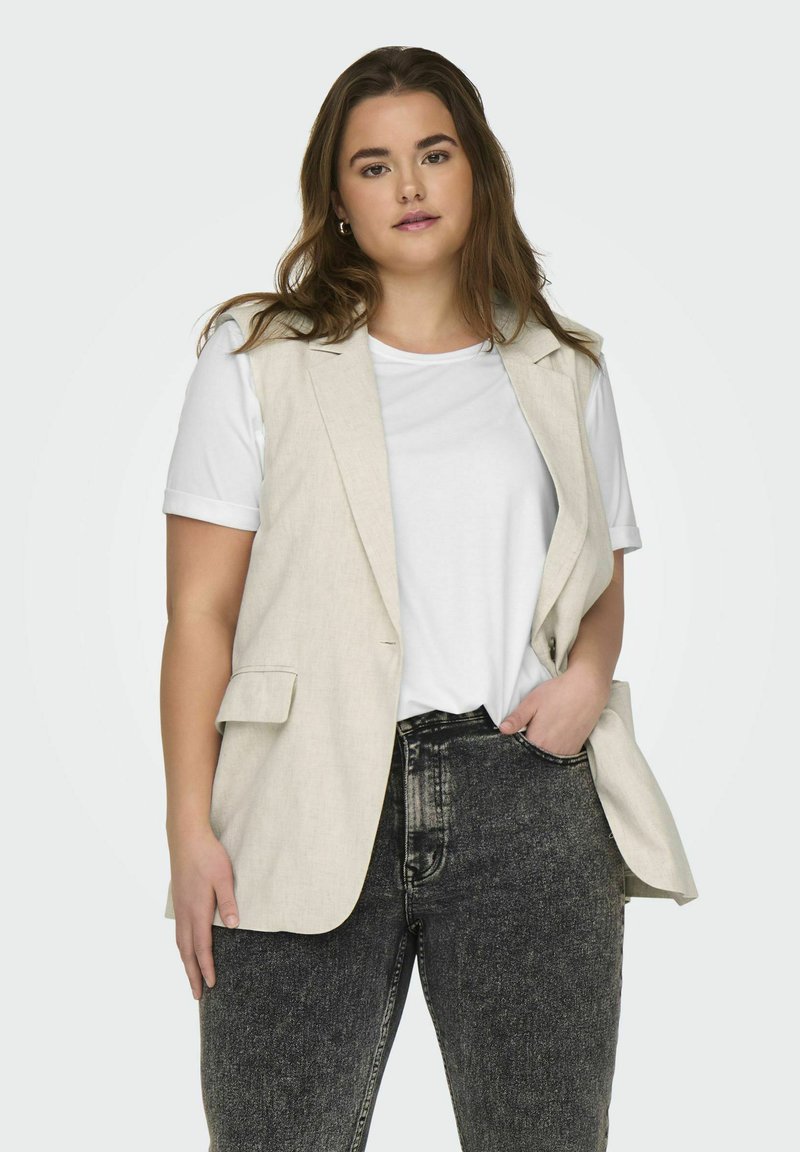 Gilet en lin beige sur un t-shirt blanc, associé à un jean en denim gris foncé. Le gilet présente des poches latérales et une coupe ajustée.