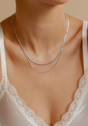 Gros plan sur le cou d’une femme portant deux délicates chaînes en argent et un top caraco blanc bordé de dentelle.